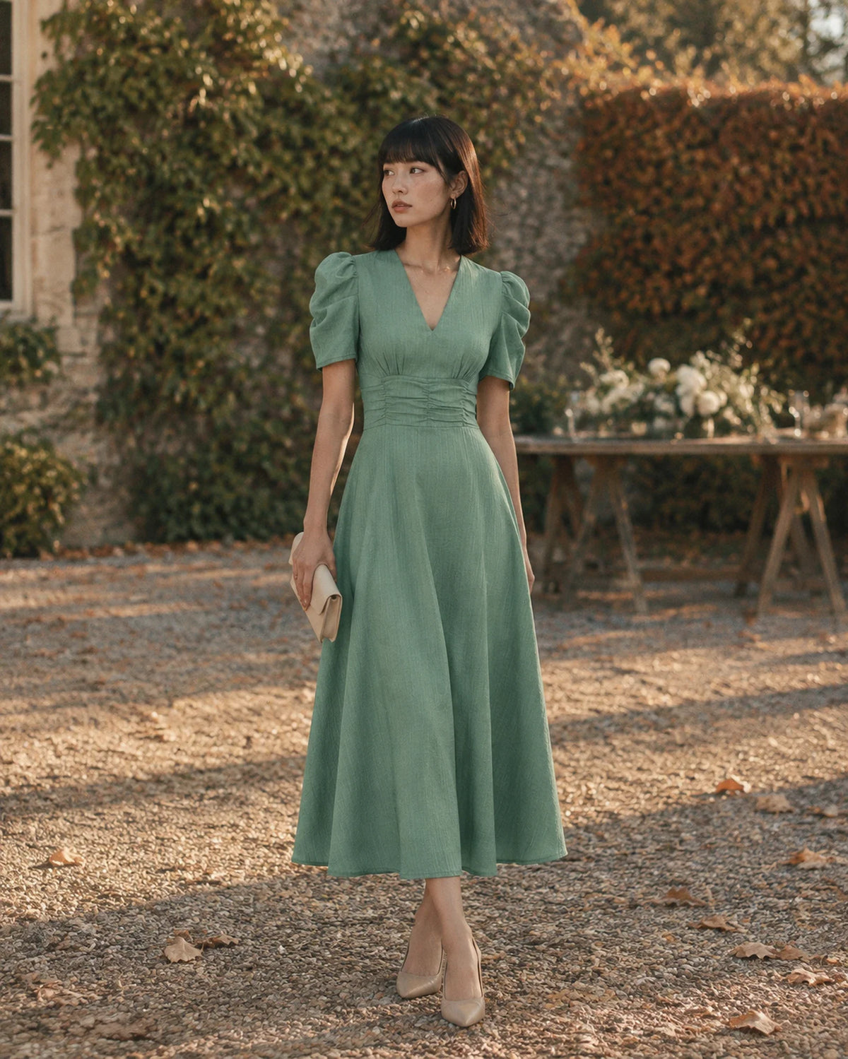 Woman in a sage green midi dress with puff sleeves standing on a gravel courtyard at an autumn wedding venue