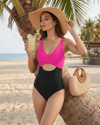 Model wearing Calypso One-Piece Swimsuit in pink and black, enjoying a drink on the beach with a sun hat.