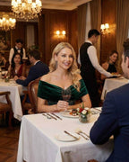 Celeste Midi Dress worn by a woman in a restaurant, featuring an elegant off-shoulder design in rich green velvet.