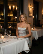 Celeste Mini Dress worn by a woman in a restaurant setting, showcasing its luxurious silver satin fabric.