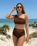 Lauven Chestnut Two-Piece Swimsuit on model at the beach, featuring a brown asymmetric top and high-waisted bottom.
