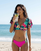 Model wearing Florence Two-Piece Swimsuit by Lauven, featuring pink floral design and tropical hues, enjoying a drink on the beach.