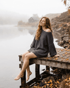 Lauven Ember Mini Dress model sitting by a lake, showcasing the cozy charcoal ribbed knit fabric and off-the-shoulder design.