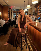 Mocha Maxi Dress by Lauven showcased on a model seated at a bar, highlighting the elegant halter neck and open back design.