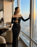 Model wearing Nocturne Maxi Dress by Lauven, off-shoulder design, holding a glass near a window with city view.