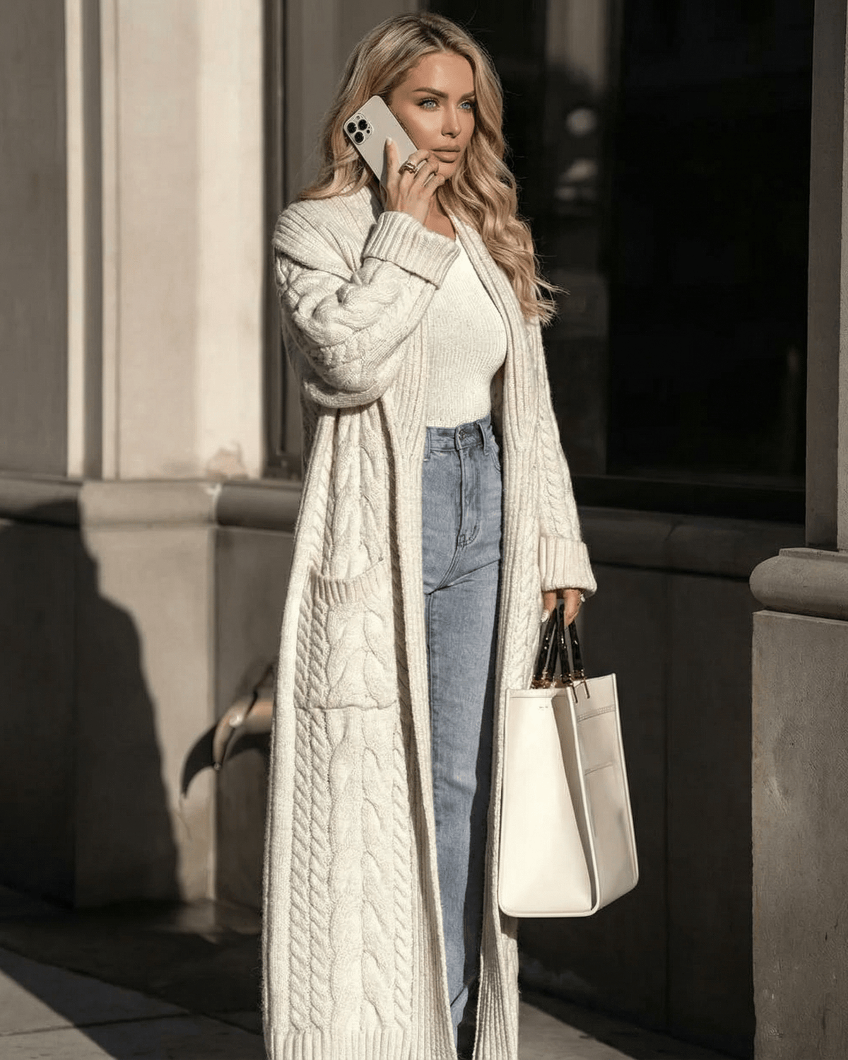 Woman wearing a Milano Cardigan while talking on a phone, paired with jeans and a handbag, captured in sunlight.