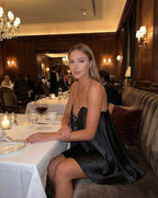 Elegant woman in Noir Mini Dress by Lauven, seated at a luxurious dining table, showcasing a satin A-line silhouette.
