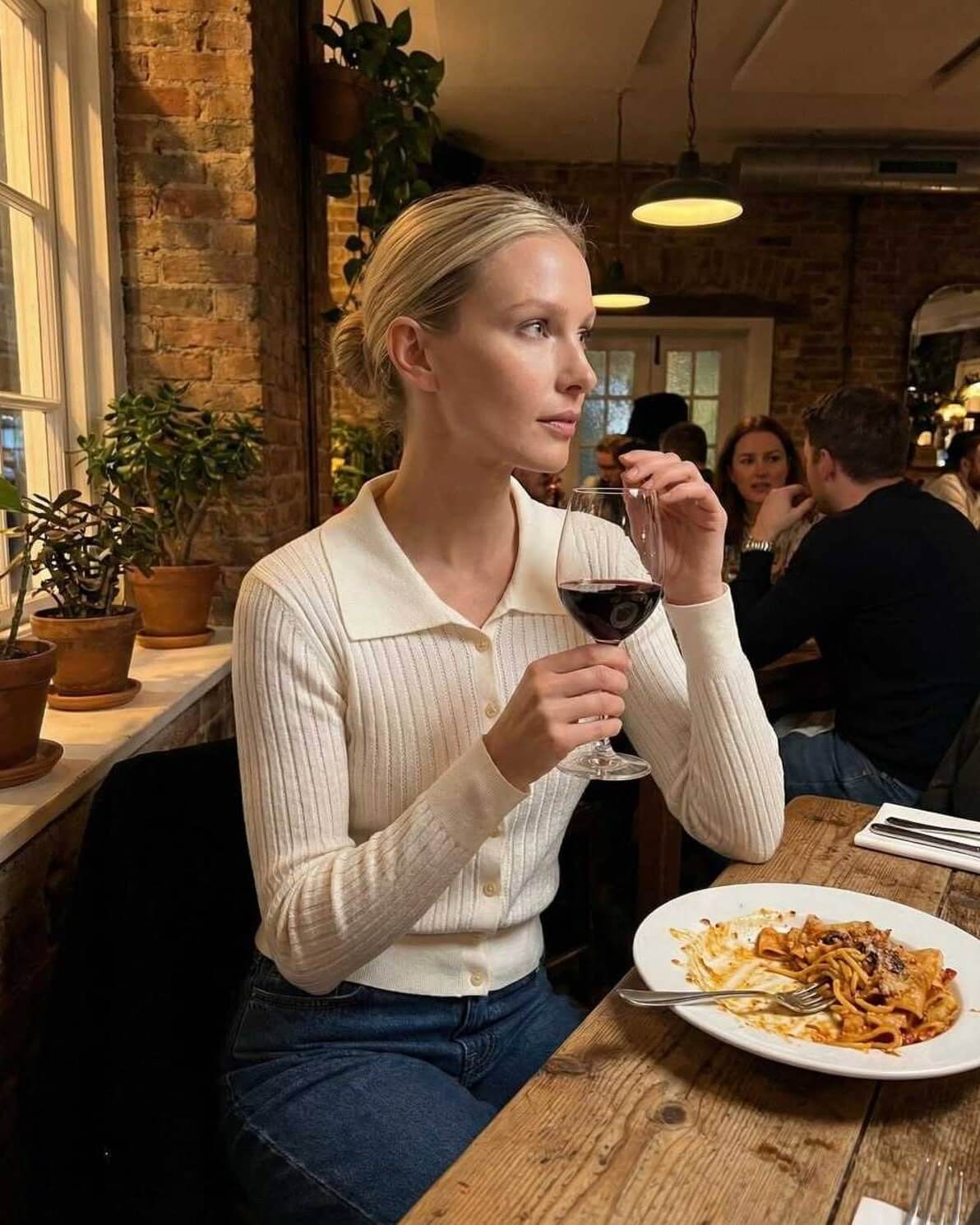 Woman in Sienna Cardigan by Lauven enjoying wine at a restaurant table with pasta.