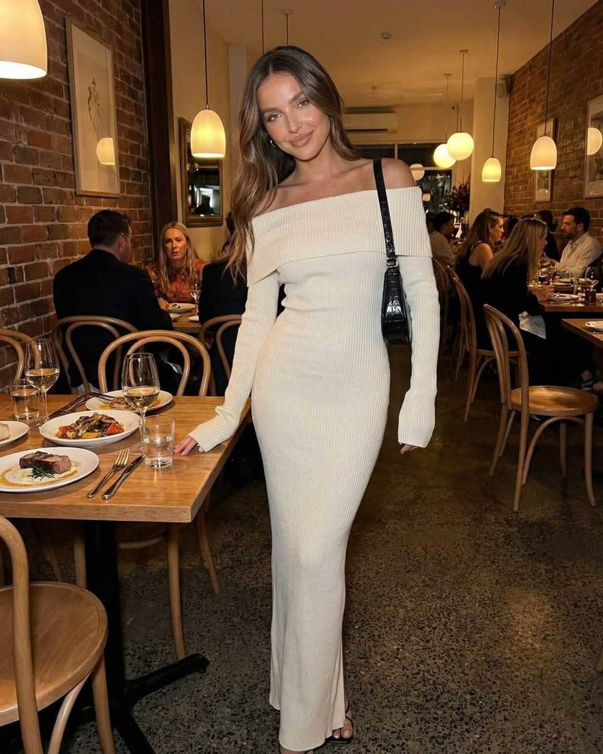 Sienna Maxi Dress by Lauven worn by a model in a restaurant, featuring an ivory off shoulder design and ribbed knit fabric.