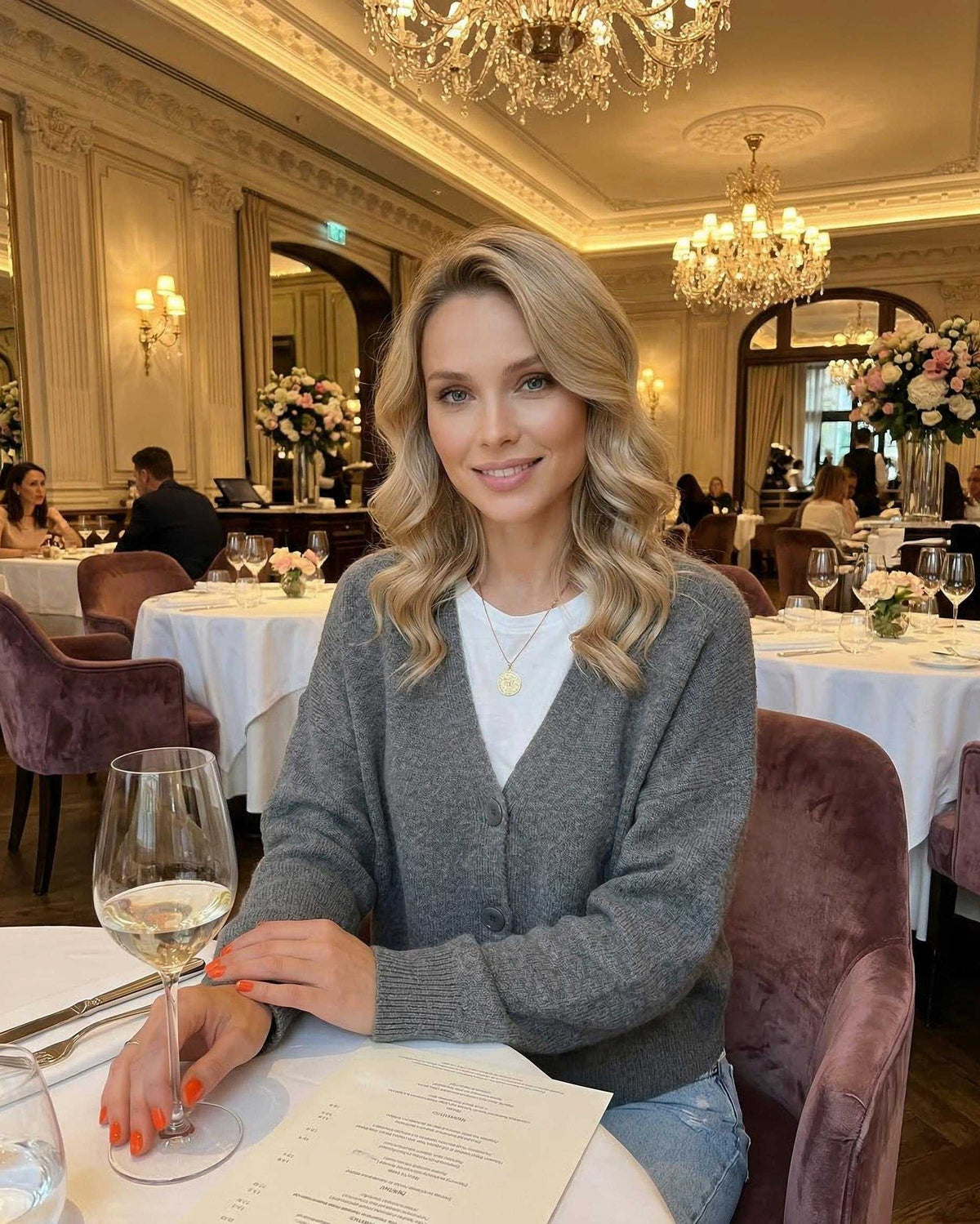 Woman wearing a Slate Cardigan by Lauven, seated at an elegant restaurant table with a glass of wine.